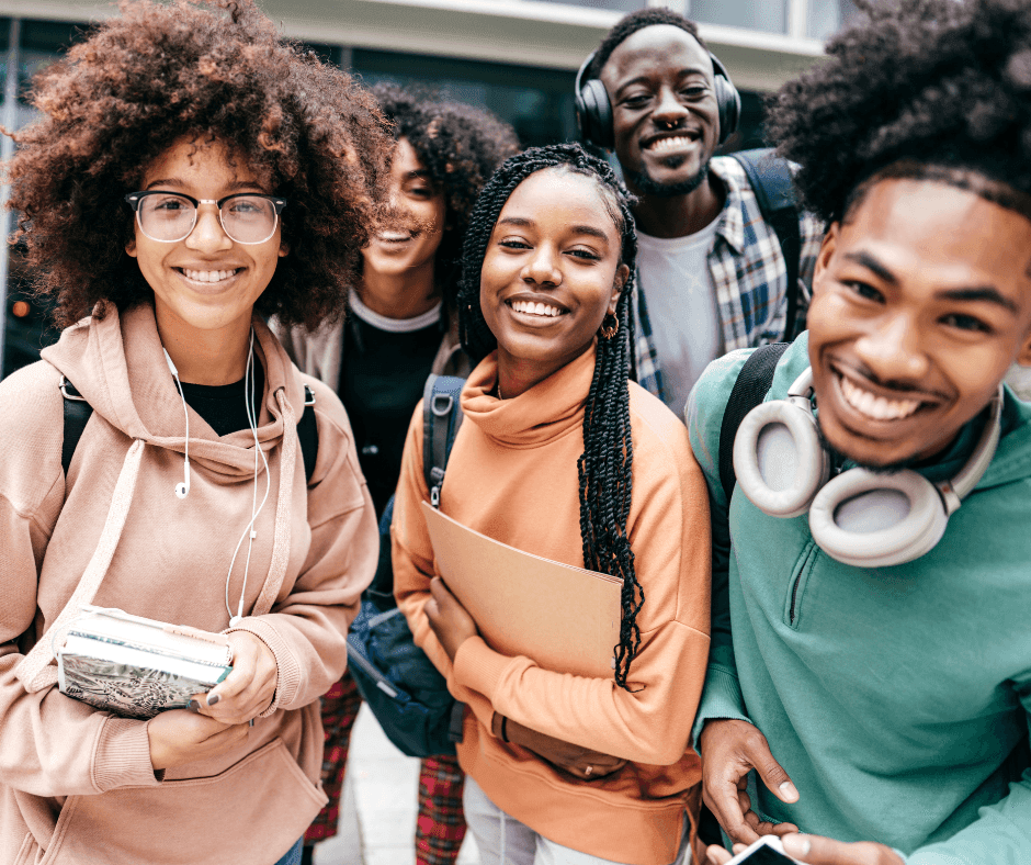 Group of individuals standing close together outdoors, wearing casual clothing and carrying books and backpacks.